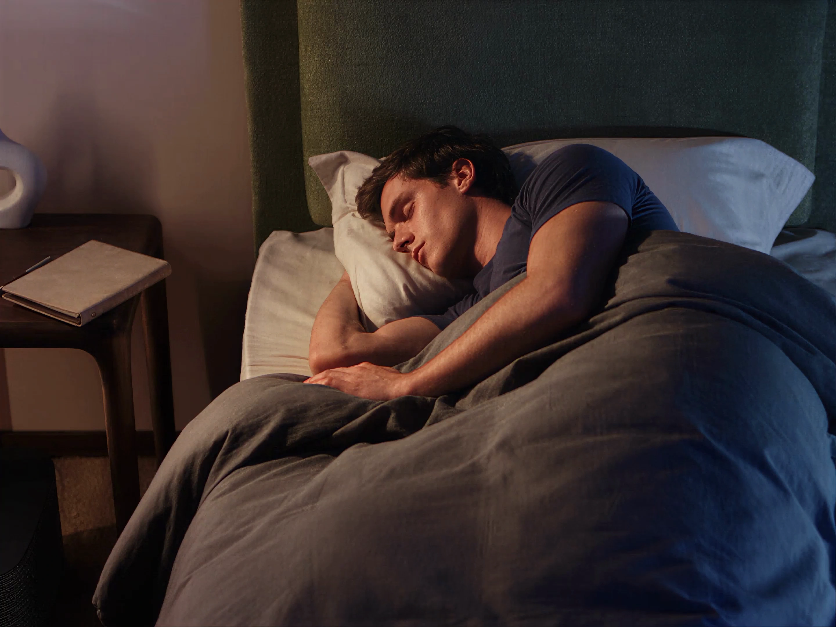 Un hombre duerme de costado en una habitación en penumbra, con la respiración moviendo suavemente la ropa de cama.