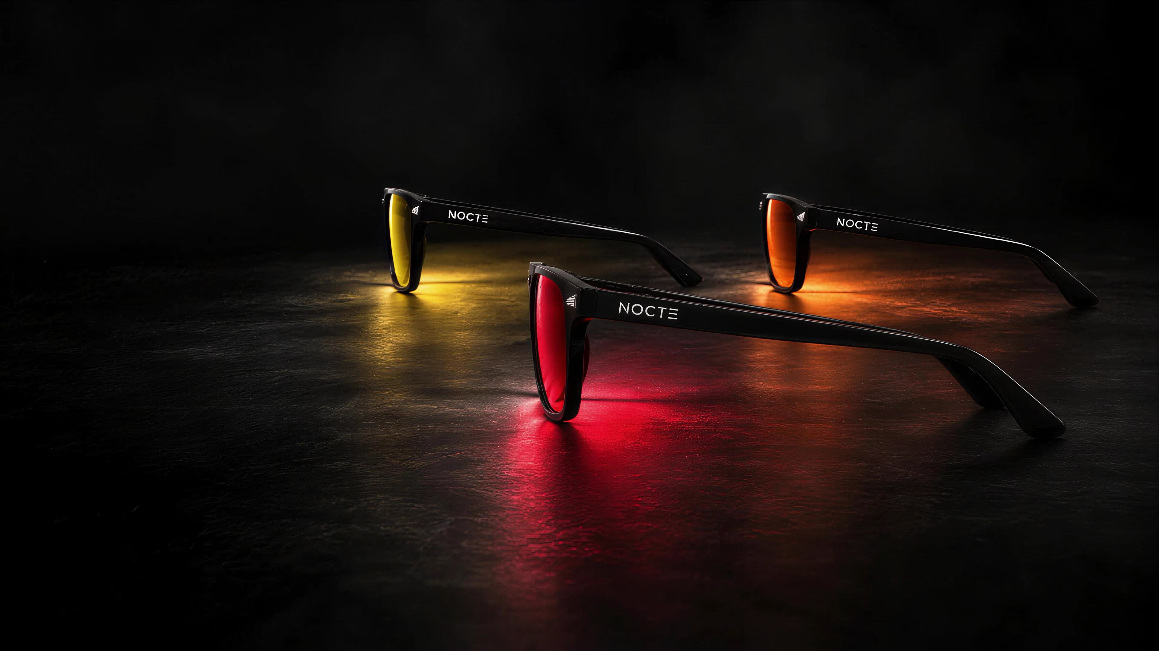 Tres lentes NOCTE de perfil: amarillo, naranja y rojo, con reflejos suaves sobre piedra oscura.