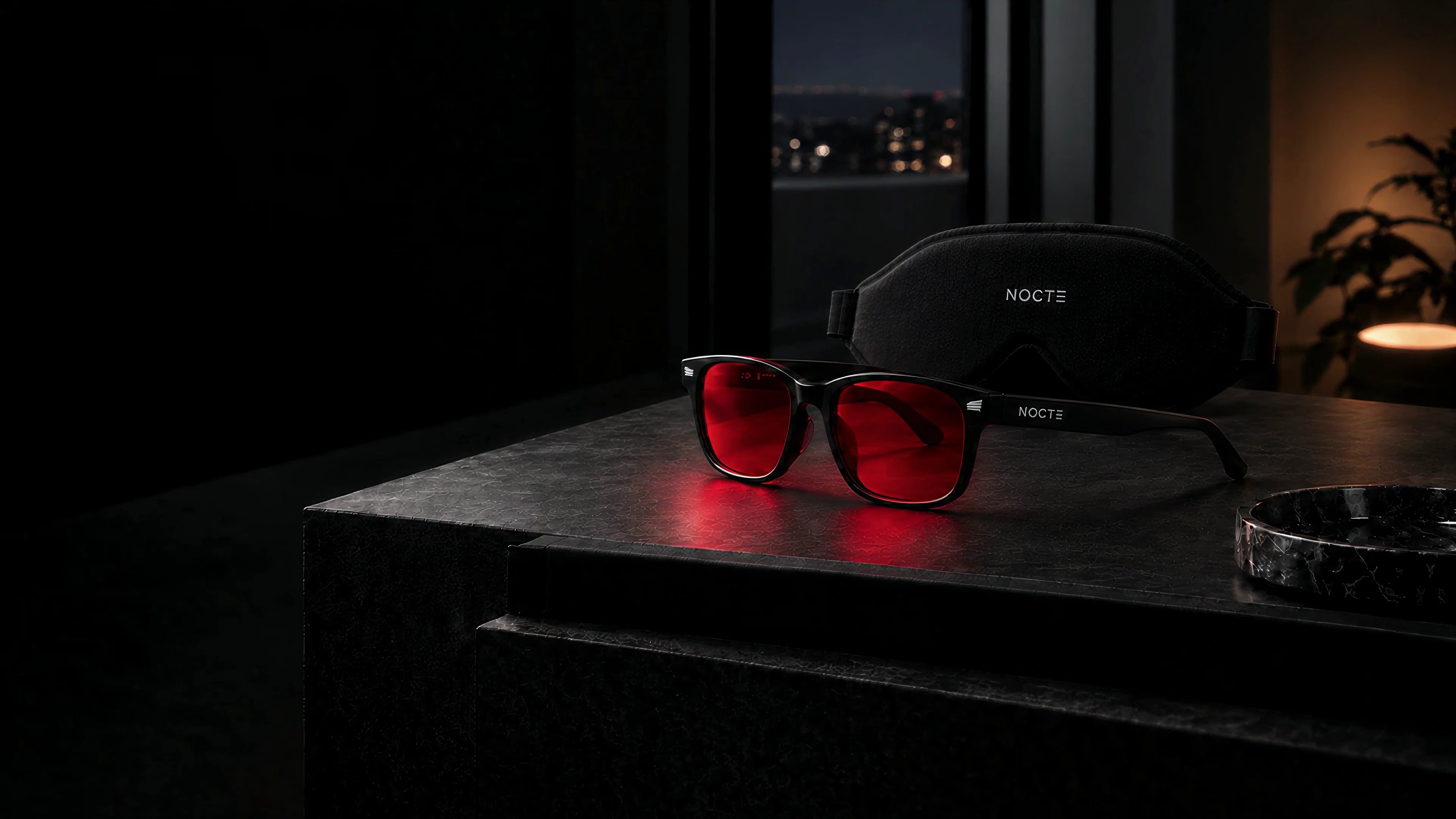 Lentes NOCTE rojos apoyados sobre piedra oscura: montura negra, detalles plateados y logo lateral.