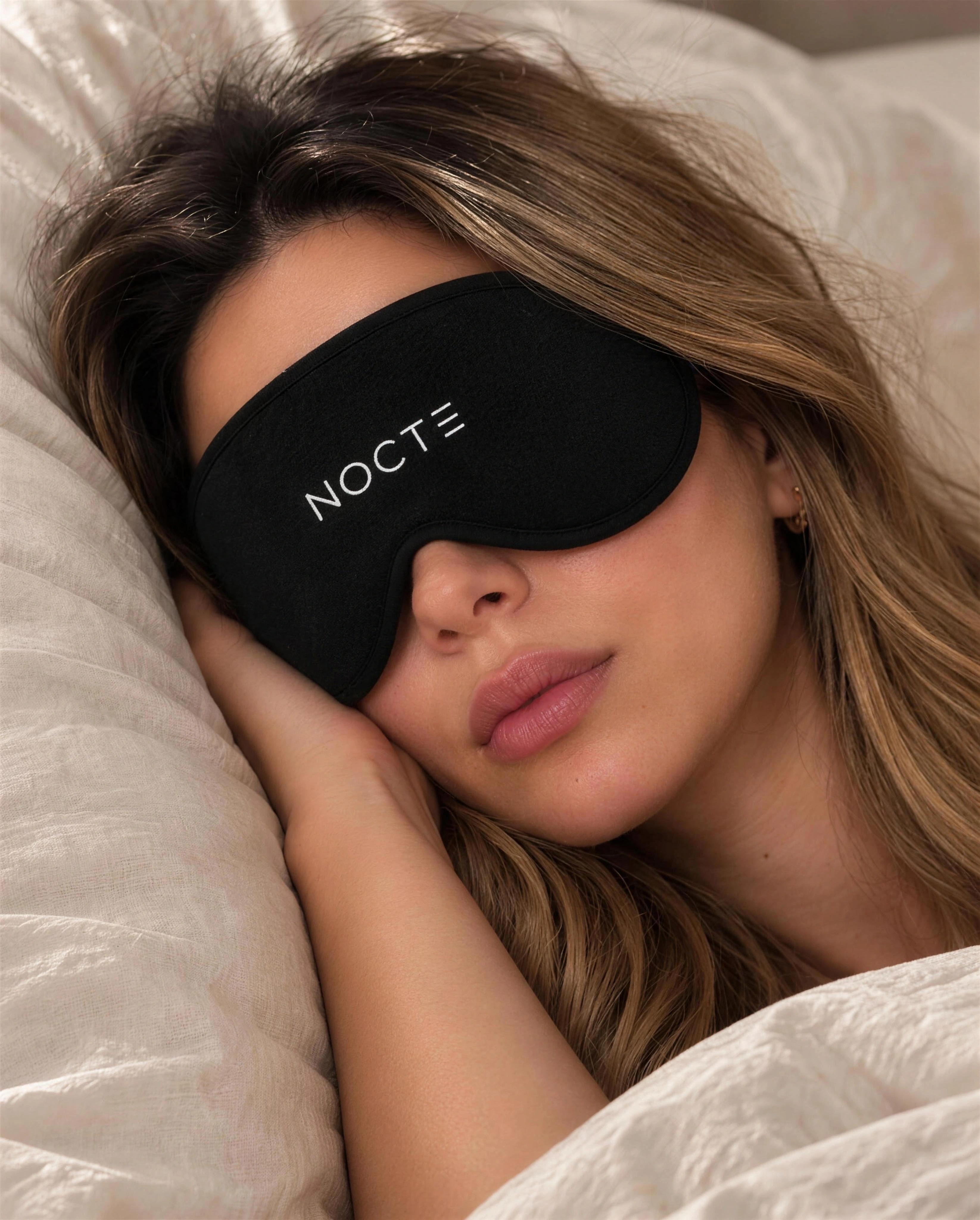 Una mujer descansa de costado sobre su almohada con el antifaz negro NOCTE.