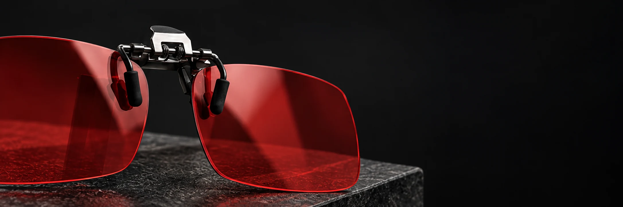 Clip-on NOCTE de lentes rojos sobre piedra oscura; se ven la pinza central y los apoyos para la montura.